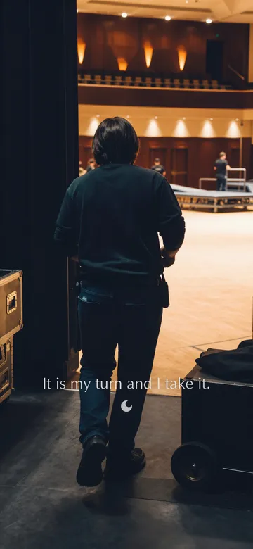 Work affirmations wallpaper: dark cinematic photograph of one person walking out of the dark wings onto a lit stage floor, seen from behind, with the affirmation 'It is my turn and I take it.' in serif type.