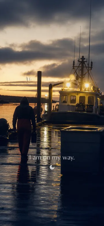 Work affirmations wallpaper: dark cinematic photograph of one person walking out along a wet jetty at dawn toward a fishing boat with its lights already on, with the affirmation 'I am on my way.' in serif type.